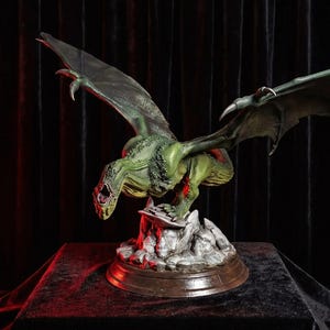 Könnte beinhalten: Eine grüne Drachenfigur mit ausgebreiteten Flügeln, scharfen Krallen und offenem Maul, die auf einem gezackten, grauen Felsensockel thront. Die Kreatur hat texturierte Schuppen und rote Akzente am Bauch und steht auf einem runden braunen Sockel.