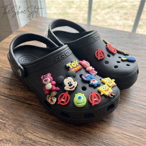 May include: A pair of black clogs decorated with various colorful character charms, including Mickey Mouse, Lotso, Stitch, Mike Wazowski, SpongeBob SquarePants, the Toy Story logo, and red Avengers logos, arranged across the top surface of the footwear.