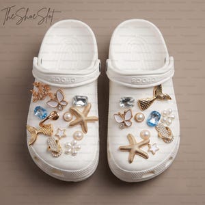 Puede incluir: Un par de zuecos blancos sin cordones con una variedad de dijes decorativos que incluyen estrellas de mar doradas, colas de sirena, mariposas blancas, gemas rectangulares azules, cristales transparentes, estrellas pequeñas y cuentas perladas.
