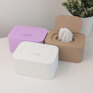 Könnte beinhalten: Drei rechteckige Aufbewahrungsboxen aus Kunststoff mit Klappdeckeln und vertikal gerippten Seiten. Eine Box ist weiß, eine hellviolett und eine beige. Die beige Box ist geöffnet und zeigt ein weißes Tuch, während die anderen geschlossen sind.