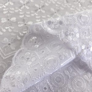 May include: Close-up of white eyelet fabric with intricate floral and geometric embroidery. The fabric is folded, revealing the detailed patterns and textures. The design features flowers, circles, and square shapes, creating a delicate and elegant look. Ideal for sewing and crafting projects.