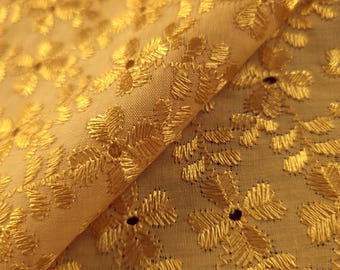 May include: Close-up of a golden fabric with embroidered floral patterns. The fabric has a soft, textured appearance with detailed stitching. The design features repeating floral motifs, creating a luxurious and elegant look. The fabric is folded, showing its depth and texture.