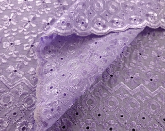 May include: Close-up of lavender eyelet fabric. The fabric features intricate floral and geometric embroidery patterns. The edges are scalloped, and the fabric is folded, showing the texture and detail of the design. This fabric is suitable for sewing projects.