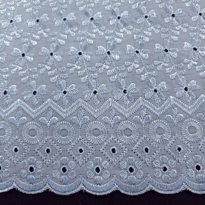 May include: Close-up of a light blue embroidered fabric with a floral and geometric pattern. The fabric features detailed stitching and small, evenly spaced holes, creating a delicate and textured appearance. The design includes flowers, circles, and diamond shapes.