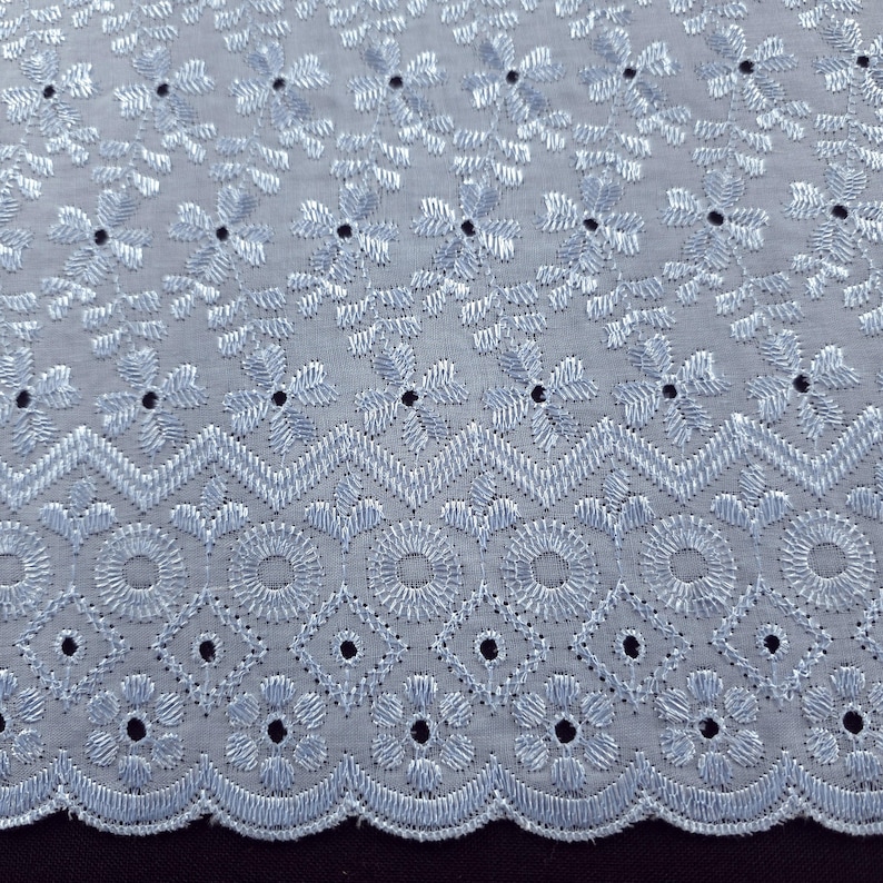 May include: Close-up of a light blue embroidered fabric with a floral and geometric pattern. The fabric features detailed stitching and small, evenly spaced holes, creating a delicate and textured appearance. The design includes flowers, circles, and diamond shapes.