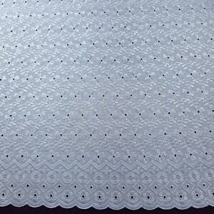 May include: Close-up of a light blue eyelet fabric. The fabric features a repeating floral pattern with small, dark blue dots. The bottom edge has a decorative scalloped border with circular and geometric cutouts. This fabric is suitable for apparel and home decor.