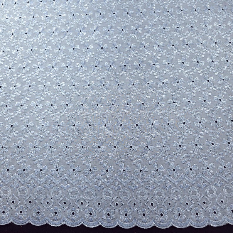 May include: Close-up of a light blue eyelet fabric. The fabric features a repeating floral pattern with small, dark blue dots. The bottom edge has a decorative scalloped border with circular and geometric cutouts. This fabric is suitable for apparel and home decor.