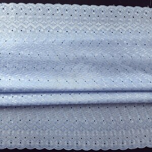 May include: Light blue eyelet fabric with a scalloped edge. The fabric features intricate floral and geometric cut-out patterns. The material is folded, showing the texture and detail of the delicate design. Ideal for sewing projects, dresses, and home decor.