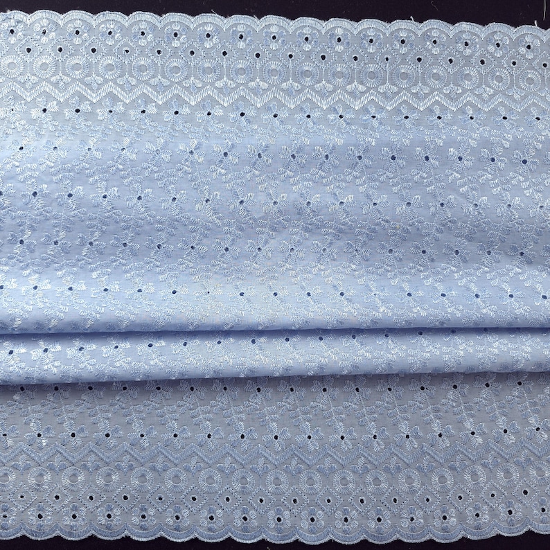 May include: Light blue eyelet fabric with a scalloped edge. The fabric features intricate floral and geometric cut-out patterns. The material is folded, showing the texture and detail of the delicate design. Ideal for sewing projects, dresses, and home decor.