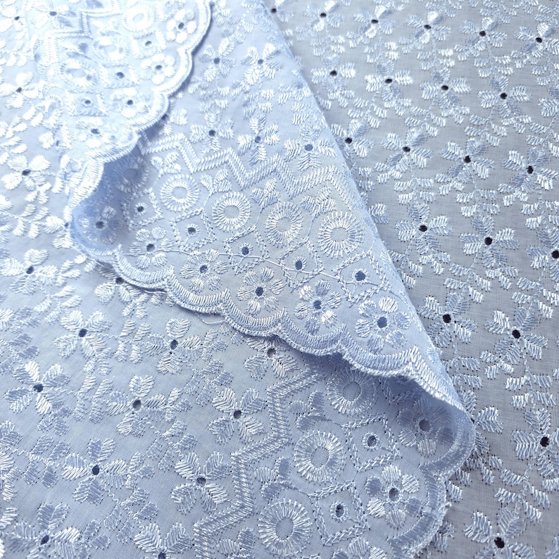May include: Close-up of light blue eyelet fabric with a scalloped edge. The fabric features intricate floral and geometric embroidery with small, evenly spaced cut-out details. The fabric is folded over, revealing the detailed pattern.