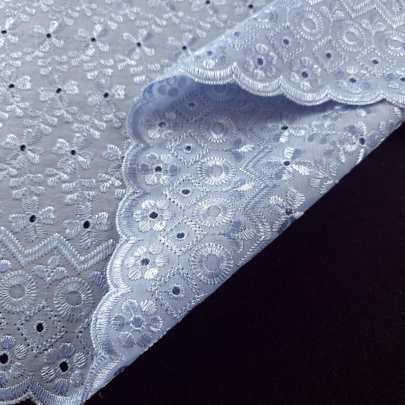 May include: Close-up of a light blue eyelet fabric. The fabric features intricate floral and geometric embroidery with scalloped edges. The fabric is folded, revealing the detailed pattern and texture.