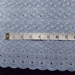 May include: Light blue embroidered fabric with a floral and geometric pattern. A measuring tape shows the fabric's width. The fabric has a scalloped edge and is suitable for various sewing projects.