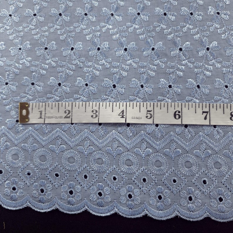 May include: Light blue embroidered fabric with a floral and geometric pattern. A measuring tape shows the fabric's width. The fabric has a scalloped edge and is suitable for various sewing projects.