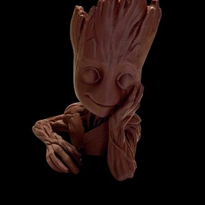 Può includere: Scultura marrone di un personaggio con testa e braccia simili a un albero, che poggia la testa sulla mano. La scultura ha un volto sorridente ed è ambientata su uno sfondo nero.