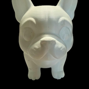Op de afbeelding: Een witte, driedimensionale sculptuur van een Franse Bulldog. De hond heeft grote, rechtopstaande oren, een korte snuit en een licht gefronste wenkbrauw. De sculptuur staat tegen een zwarte achtergrond.