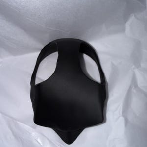 Op de afbeelding: Een zwart, gevormd gezichtsmasker met een gebogen bovenkant en een puntige onderkant. Het masker heeft twee grote openingen voor de ogen en een band die over het hoofd gaat. Het masker is gemaakt van een glad, mat materiaal.