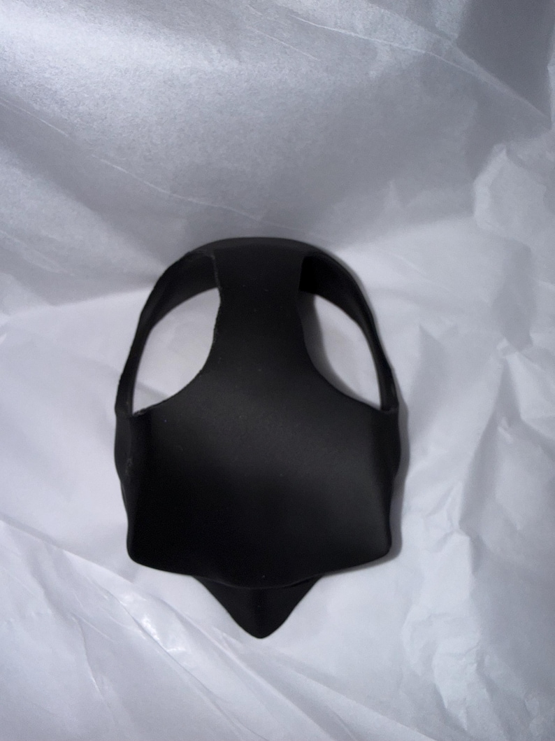 Op de afbeelding: Een zwart, gevormd gezichtsmasker met een gebogen bovenkant en een puntige onderkant. Het masker heeft twee grote openingen voor de ogen en een band die over het hoofd gaat. Het masker is gemaakt van een glad, mat materiaal.