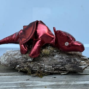 May include: A plush dragon toy crafted from a shiny, metallic red fabric. The creature features a long, pointed tail, folded wings, and a single white button serving as an eye. The stuffed animal is positioned in a resting pose with its head lowered.