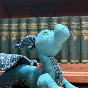 May include: A teal plush dragon toy featuring dark blue curved horns with stripes, a smooth fabric body, and textured, speckled blue and white fabric wings. The creature has a rounded head with a snout and bright blue eyes looking upward.