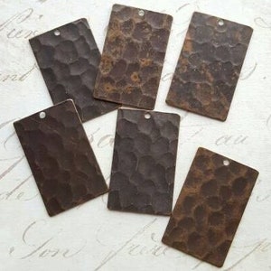 Puede incluir: Seis etiquetas rectangulares de metal con textura martillada. Las etiquetas son de color marrón oscuro y tienen un agujero en la parte superior para colgar.