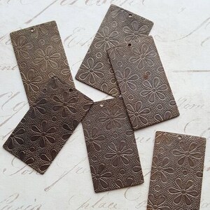 May include: Six brown metal tags with a floral pattern. The tags have holes punched in the top for hanging.