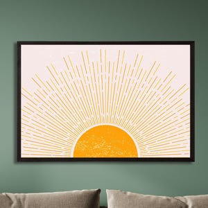 May include: A rectangular wall art piece in a black frame featuring a graphic illustration of a bright orange sun rising from the bottom center. The sun has a textured surface and emits thin, radiating yellow lines against a pale pink background.