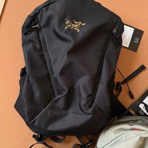 May include: Black backpack with a gold logo on the front. The backpack has a top handle, two zippered pockets, and adjustable straps. A tag is attached to the side. The backpack is made of a durable, water-resistant material.