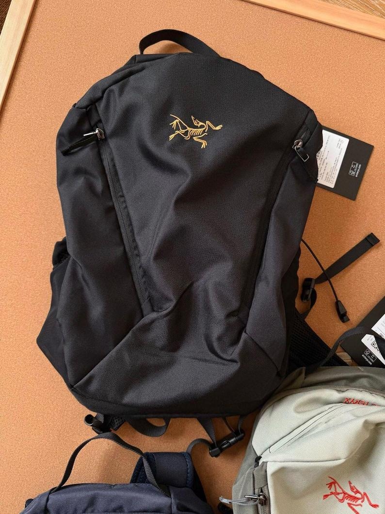 May include: Black backpack with a gold logo on the front. The backpack has a top handle, two zippered pockets, and adjustable straps. A tag is attached to the side. The backpack is made of a durable, water-resistant material.