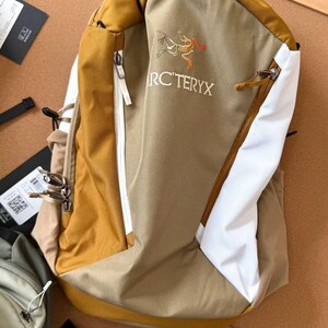 May include: A tan and white backpack with a mustard yellow top and accents. The backpack has a zippered front pocket and the text "ARC'TERYX" is visible. The backpack is made of a durable material.