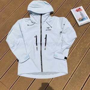 May include: A light blue hooded jacket with a full-length zipper and two front pockets. The jacket has the brand name "Arc'teryx" printed on the chest. The jacket is on a wooden surface.