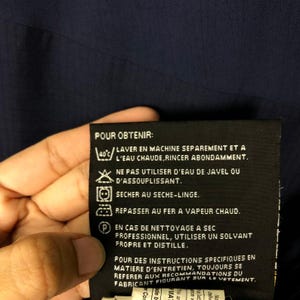 Puede incluir: Una etiqueta de cuidado negra con texto y s&iacute;mbolos blancos, sostenida por una mano. La etiqueta incluye instrucciones de lavado, como lavar a m&aacute;quina por separado a 40&deg;C, no usar lej&iacute;a y secar en secadora. Tambi&eacute;n tiene instrucciones de planchado y limpieza en seco.