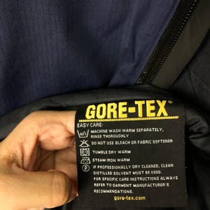 Puede incluir: Primer plano de una etiqueta negra GORE-TEX con texto amarillo e instrucciones de cuidado. La etiqueta est&aacute; adherida a una prenda azul marino. El texto incluye instrucciones de lavado y el sitio web gore-tex.com.