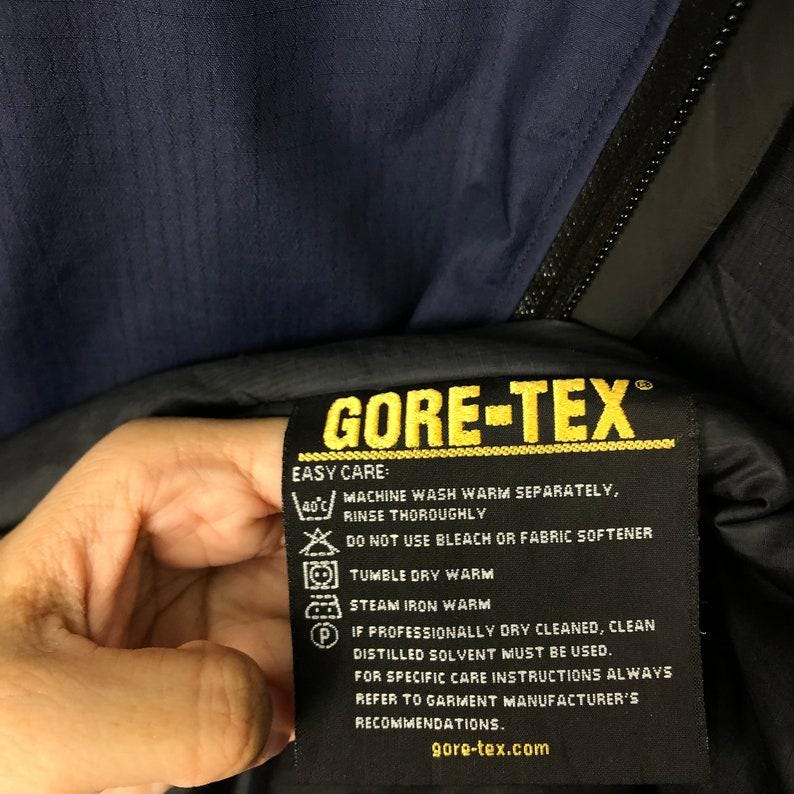Puede incluir: Primer plano de una etiqueta negra GORE-TEX con texto amarillo e instrucciones de cuidado. La etiqueta est&aacute; adherida a una prenda azul marino. El texto incluye instrucciones de lavado y el sitio web gore-tex.com.