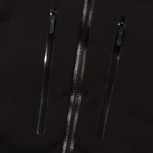 May include: Close-up of a black garment featuring three zippers. Two long, thin zippers are positioned on either side of a central zipper. The central zipper has a silver-colored zipper pull.