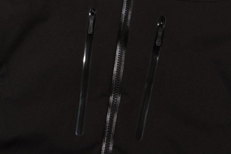 May include: Close-up of a black garment featuring three zippers. Two long, thin zippers are positioned on either side of a central zipper. The central zipper has a silver-colored zipper pull.