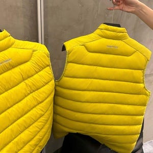 May include: Two bright yellow puffer vests with horizontal stitching are displayed. The vests are sleeveless with a high collar and a small logo on the back. One vest is held by a hanger, and the other is partially visible on the left.