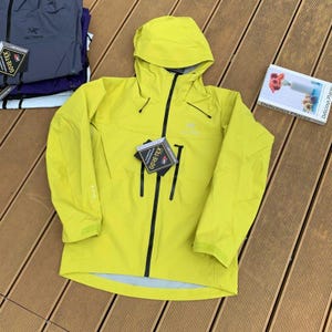 May include: A bright yellow hooded jacket with a black zipper and black accents. The jacket has a Gore-Tex tag attached. Additional jackets are visible in the background.
