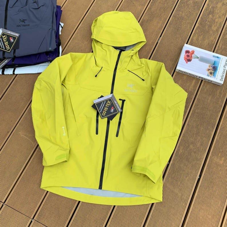 May include: A bright yellow hooded jacket with a black zipper and black accents. The jacket has a Gore-Tex tag attached. Additional jackets are visible in the background.