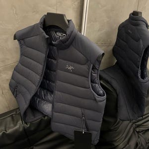 May include: Two dark gray puffer vests hanging on hangers. The vests have a quilted design and a small logo on the chest. One vest is open, revealing a black interior. A black tag hangs from one of the vests.