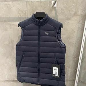 May include: Navy blue puffer vest with a high collar and a full-length zipper. The vest has two zippered pockets and a small logo on the chest. A tag hangs from the zipper. The vest is hanging on a black hanger.