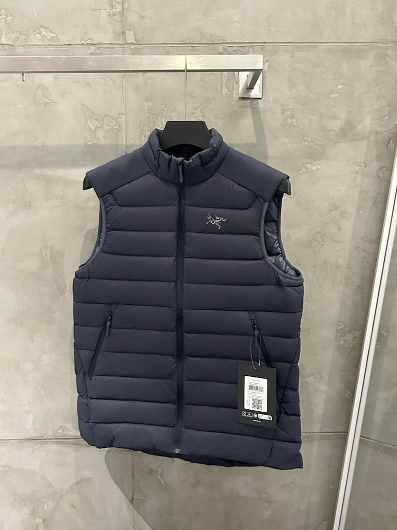 May include: Navy blue puffer vest with a high collar and a full-length zipper. The vest has two zippered pockets and a small logo on the chest. A tag hangs from the zipper. The vest is hanging on a black hanger.
