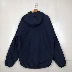 Puede incluir: Una chaqueta con capucha azul marino colgada de una percha de madera. La chaqueta tiene un forro negro y pu&ntilde;os negros. La capucha est&aacute; levantada y la chaqueta se ve desde atr&aacute;s. La chaqueta parece estar hecha de un material liso, posiblemente resistente al agua.