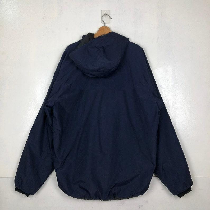 Puede incluir: Una chaqueta con capucha azul marino colgada de una percha de madera. La chaqueta tiene un forro negro y pu&ntilde;os negros. La capucha est&aacute; levantada y la chaqueta se ve desde atr&aacute;s. La chaqueta parece estar hecha de un material liso, posiblemente resistente al agua.