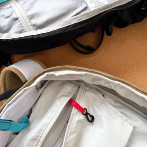 May include: Close-up of two open backpacks. One is black with a gray interior and a teal strap. The other is beige with a white interior and a red zipper pull. Both have zippers and black hardware.