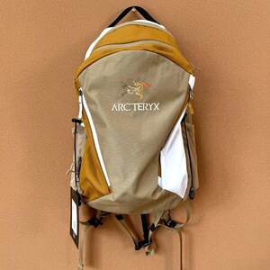 May include: A tan and mustard-colored backpack with the Arc'teryx logo. The backpack has white accents, a black handle, and adjustable straps. It features multiple compartments and a zipper closure. A tag hangs from the bottom.