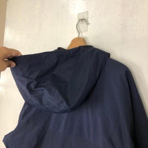Puede incluir: Chaqueta con capucha azul marino colgada de una percha de madera. La chaqueta es de un material liso, posiblemente resistente al agua. La capucha es prominente y la chaqueta parece estar en buen estado.