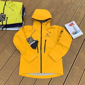 May include: Bright yellow hooded jacket with a black zipper and pockets. The jacket has the brand name "Arc'teryx" and a "Gore-Tex" tag. A stack of similar jackets in yellow and purple are visible in the background. A book titled "Delicious Places" is also visible.