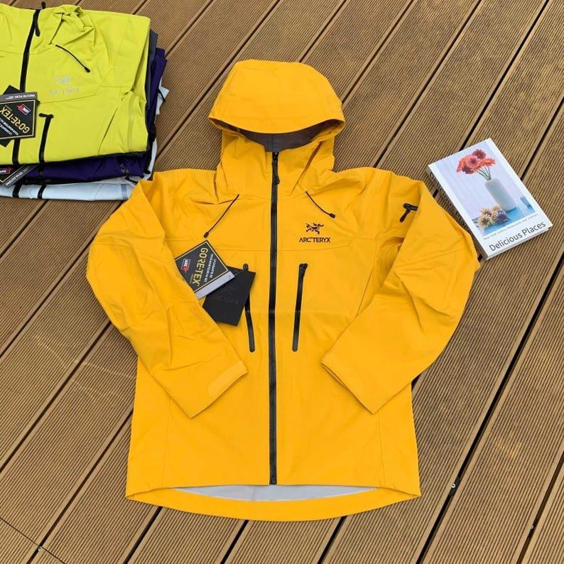 May include: Bright yellow hooded jacket with a black zipper and pockets. The jacket has the brand name "Arc'teryx" and a "Gore-Tex" tag. A stack of similar jackets in yellow and purple are visible in the background. A book titled "Delicious Places" is also visible.