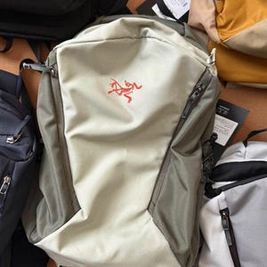 May include: A light beige and gray backpack with a red logo on the front. The backpack has two zippered compartments and a drawstring closure. Several other backpacks are visible in the background.