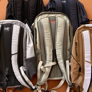 May include: A collection of backpacks in various colors, including black, navy, gray, and tan. Each backpack features a padded back panel, adjustable straps, and a logo. The backpacks are designed for carrying items.